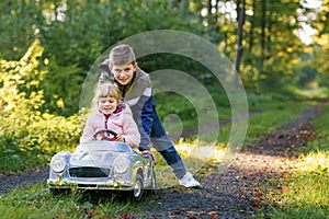 Two happy children playing with big old toy car in autumn forest, outdoors. Kid boy pushing and driving car with little