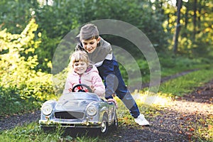 Two happy children playing with big old toy car in autumn forest, outdoors. Kid boy pushing and driving car with little