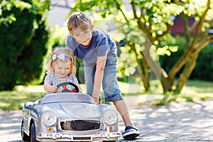 Two happy children playing with big old toy car in summer garden, outdoors. Boy driving car with little girl, cute sister inside.