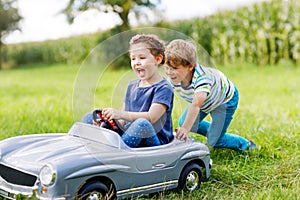 Two happy children playing with big old toy car in summer garden, outdoors. Boy driving car with little girl inside