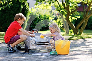 Two happy children washing big old toy car in summer garden, outdoors. Brother boy and little sister toddler girl