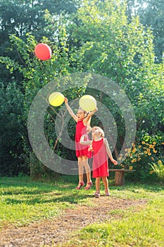 Two happy cute little sisters jumping with colorful toy balloons outdoors. Smiling kids having fun in green spring garden at warm