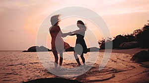 Two happy dancing girls holding hands on the beach at sunset in slow motion