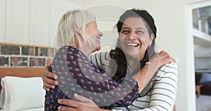 Two happy diverse senior women embracing and laughing in sunny bedroom, slow motion