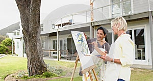 Two happy diverse senior women painting picture and laughing in sunny garden, slow motion