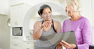 Two happy diverse senior women using tablet and laughing in kitchen, slow motion