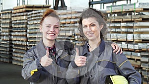 Two happy female factory workers embracing showing thumbs up at the storage