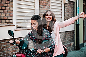 Two happy female friends riding on motorbike together through city street