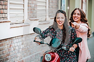 Two happy female friends riding on motorbike together through city street, while young woman pointing finger away