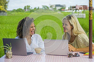 Two happy female friends working outdoors at beautiful internet cafe with laptop computer caucasian woman and an afro mixed girl