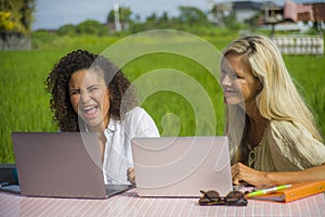 Two happy female friends working outdoors at beautiful internet cafe with laptop computer caucasian woman and an afro mixed girl