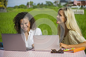 Two happy female friends working outdoors at beautiful internet cafe with laptop computer caucasian woman and an afro mixed girl