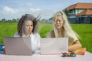 Two happy female friends working outdoors at beautiful internet cafe with laptop computer caucasian woman and an afro mixed girl