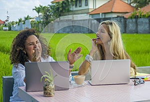 Two happy female friends working outdoors at beautiful internet cafe with laptop computer caucasian woman and an afro mixed girl