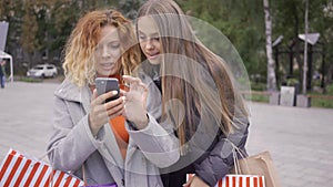 Two happy friends on the street looking at the mobile phone. Couple of fashion women with shopping bags in the city