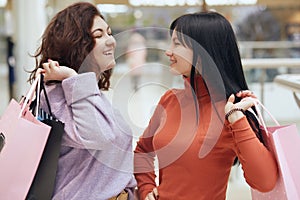 Two happy girlfriends looking smiling at each other while standing with shopping bags in mall, ladies wearing casual attire, being