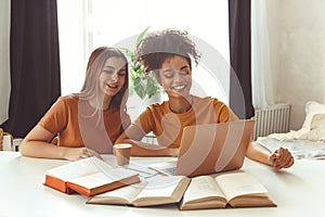 Two happy girlfriends sitting in front of laptop computer, preparing for exams together at home