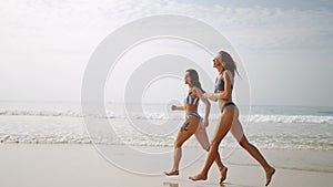 Two happy girls in bikini running at sea water waves. Cheerful female friends having fun, tanning on summer day at