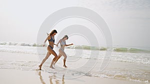 Two happy girls in bikini running at sea water waves. Cheerful female friends having fun, tanning on summer day at