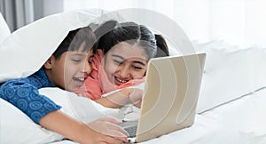 Two happy Indian brother and sister in traditional clothing lying on bed under blanket smiling, using laptop, having fun together