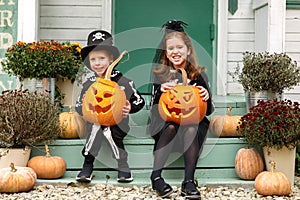 Two happy kids in Halloween costumes sitting on porch of their house with carved pumpkins