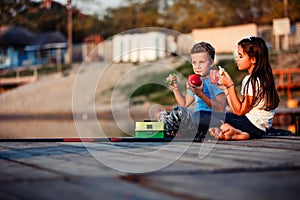 Two happy little friends, boy and girl talking, drinking tea, eating sandwiches and fishing on a lake in a sunny summer day