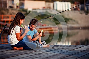 Two happy little friends, boy and girl talking, eating sandwiches and fishing on a lake