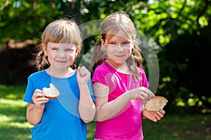 Two happy little girls eating slices of bread with butter smiling, outdoors portrait. Kids, sisters, children eating food outside