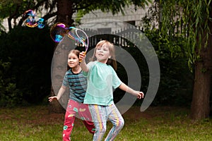 Two happy little girls running around playing with soap bubbles in the backyard. Active healthy young children making bubbles