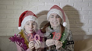 Two happy little girls show the camera figures 2019. They sit and dance in Santa`s hats on the couch against a white