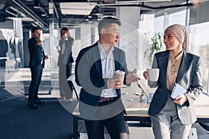 Two happy multiracial colleagues talking and drinking coffee during break time in office