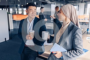 Two happy multiracial colleagues talking and drinking coffee during break time in office