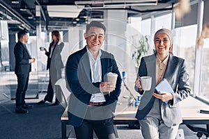 Two happy multiracial colleagues talking and drinking coffee during break time in office