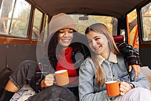 Two happy multiracial girlfriends on car trunk in autumn forest, embracing and drinking hot tea