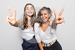 Two happy pretty women posing together and showing peace gestures while looking at the camera over gray background