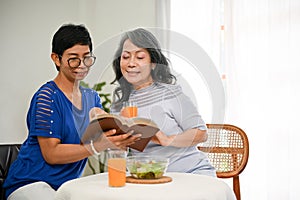 Two happy retired female are discussing something in a book while enjoying their healthy brunch