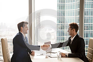 Two happy satisfied businessmen handshaking in office, city at b