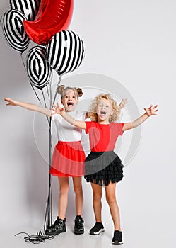 Two happy screaming singing kids girls in skirts and t-shirts with air balloons stand together with arms outstretched