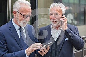 Two happy senior businessmen making phone calls, standing on the sidewalk