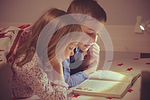 Two happy sibling children reading book in bunk bed under blanket