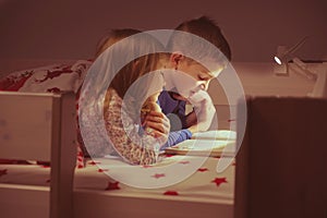 Two happy sibling children reading book in bunk bed under blanket