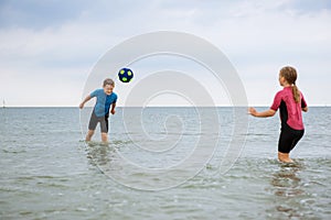 Two happy siblings children playing and jumping with ball in water in neoprene suits in sea