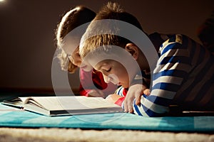 Two happy siblings reading book at darkness with flashlight at h