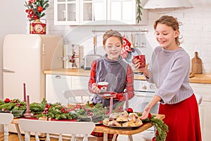 Two happy sisters are decorating the Christmas table home in kitchen, holding cups and sweet pies in their hands