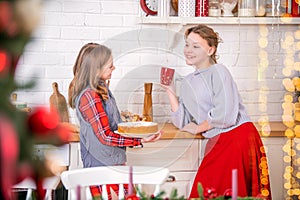 Two happy sisters are decorating the Christmas table home in kitchen, holding cups and sweet pies in their hands