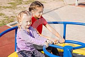 Two happy smiling little girls having fun on playground, riding on a merry-go-round