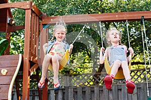 Two happy smiling little girls on swing on backyard playground outside on summer day