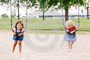 Two happy smiling little toddlers girls friends swinging on swings at playground outside