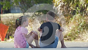 Two happy smiling teenage children, boy and girl sitting outdoors resting having fun on summer sunny day