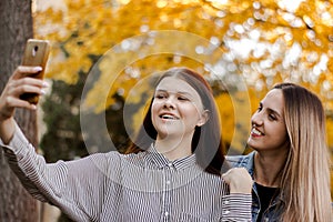 Two happy smiling young girls taking selfie on the phone in the autumn park